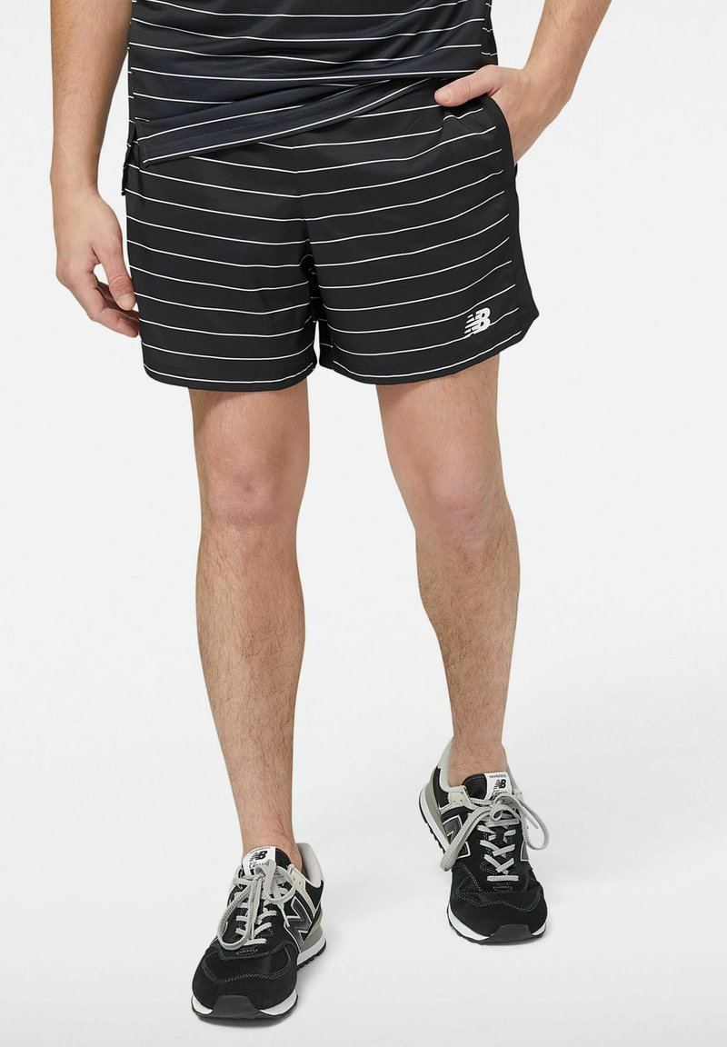 Schwarz-weiß gestreifte Sportshorts mit glatter Textur, ausgestattet mit einem elastischen Bund und seitlichen Taschen, kombiniert mit schwarzen Turnschuhen.