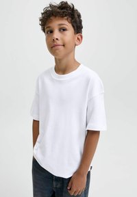 Garçon aux cheveux bouclés portant un t-shirt blanc surdimensionné et un jean noir, se tenant les mains dans les poches, regardant légèrement sur le côté.