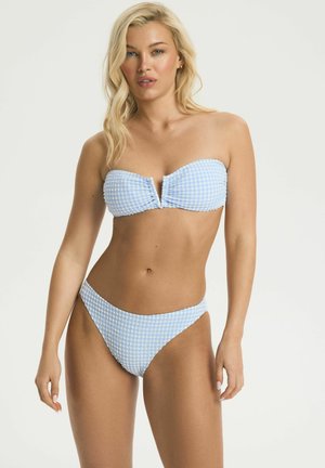 Blonde woman wearing a light blue and white checkered strapless bikini, standing against a plain light background.