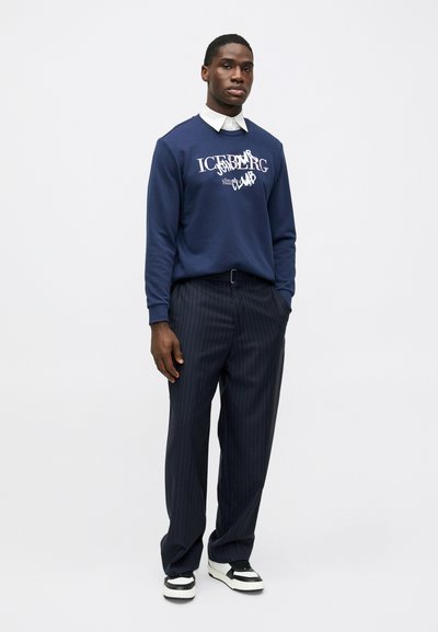 Ναυτικό φούτερ με λευκό λογότυπο, συνδυασμένο με ρασέλ παντελόνι και μαύρα-άσπρα αθλητικά παπούτσια. Χαλαρή εφαρμογή και απαλές υφές.