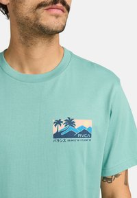 Turquoise t-shirt met korte mouwen, voorzien van een grafische patch met palmbomen, bergen, zonsondergang en tekst. Gemaakt van zachte katoenen stof.
