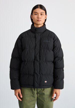 Giacca a puffo nera con colletto alto, design trapuntato e logo sul petto. Indossata sopra pantaloni verde oliva, dotata di tasche laterali.