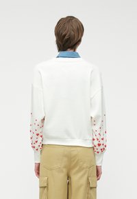 Balta sporta džemperis ar apaļu kakla izgriezumu un garām piedurknēm, ar izkaisītām sarkanu sirdi rakstiem uz piedurknēm. Apvienots ar bēšām kravas biksēm.