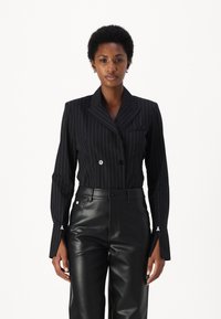 Blazer nero a doppio petto con righe verticali, collo a punta, vestibilità su misura e cuciture bianche decorative sui polsini; abbinato a pantaloni in pelle nera.
