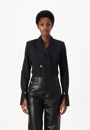 Blazer nero a doppio petto con righe verticali, collo a punta, vestibilità su misura e cuciture bianche decorative sui polsini; abbinato a pantaloni in pelle nera.
