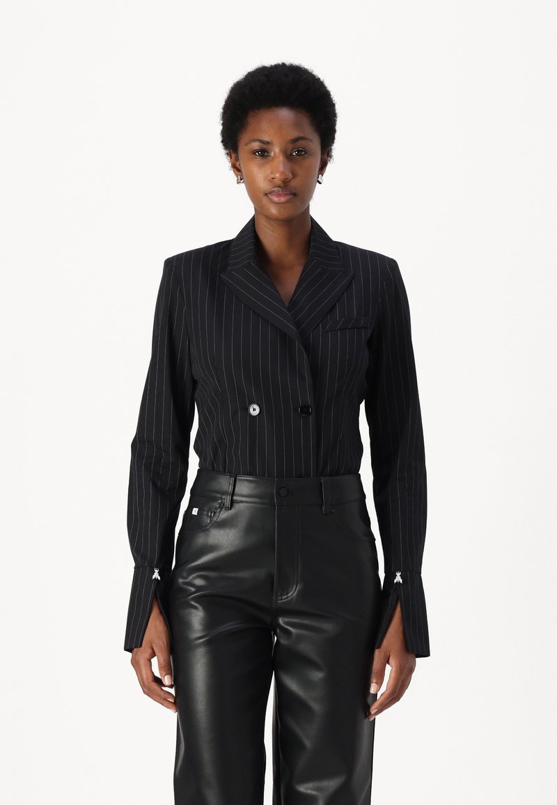Blazer nero a doppio petto con righe verticali, collo a punta, vestibilità su misura e cuciture bianche decorative sui polsini; abbinato a pantaloni in pelle nera.