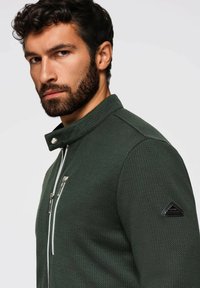 Mörkgrön jacka med en texturerad yta, med hög krage, silverfärgad dragkedja och en svart triangulär logotyp på ärmen.