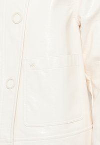 Κοντινό πλάνο από τσέπη λευκού τζάκετ με λογότυπο "AX" και μεγάλους κουμπιά σνάπ σε υφασμάτινη υφή.