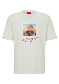 Camiseta blanca de algodón con un gran estampado gráfico de una montaña y un animal desenfocado. El texto "Hugs" en cursiva roja se encuentra debajo de la imagen.