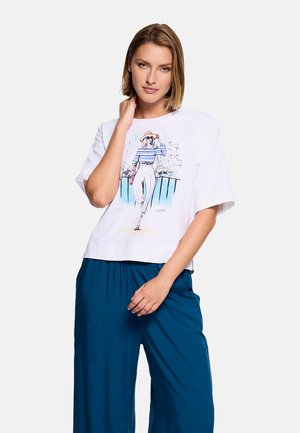 Mujer con camiseta blanca de manga corta con una ilustración colorida de una mujer con sombrero y camiseta de rayas, combinada con pantalones azules holgados.