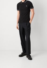 Černé polo tričko s bílým límečkem a akcenty na rukávech, logo CK na hrudi. S kombinovanými černými kalhotami na míru a černými botami.