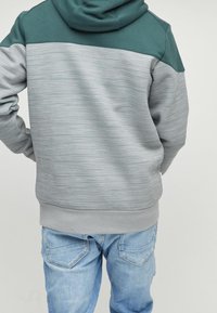 Osoba w niebiesko-szarym paskowanym hoodie z rękami w kieszeniach i jasnoniebieskich dżinsach, odwrócona tyłem na jednolitym jasnym tle.