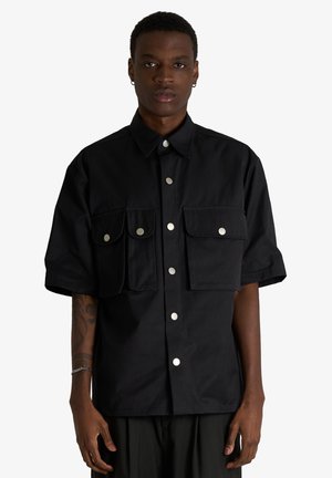 Giovane uomo che indossa una camicia nera a maniche corte larga con bottoni argentati e due grandi tasche frontali, in piedi davanti a uno sfondo semplice.