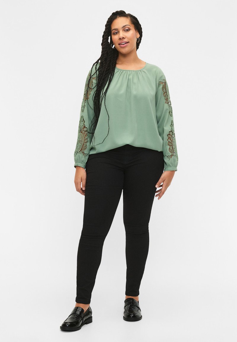 Blusa verde con maniche lunghe ricamate, scollatura rotonda, abbinata a jeans skinny neri e scarpe nere lucide. Tessuto morbido, design casual.