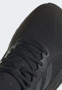 Czarne buty sportowe z teksturowaną górą z siateczki, z czarnymi sznurowadłami i trzema szarymi paskami po bokach. Wygięty kształt i wyściełany kołnierz.