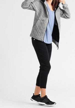 Giacca atletica grigia con cappuccio, pantaloni neri con zip e sneakers nere. Camicia blu sotto; presenta un tessuto testurizzato e tasche laterali.
