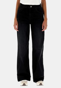 Persona che indossa jeans neri a vita alta e a gamba larga, abbinati a sneakers bianche, in piedi contro uno sfondo neutro.