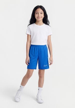 Młoda dziewczyna stoi, ubrana w białą koszulkę, niebieskie szorty Nike, białe skarpetki Nike oraz białe sportowe buty, na jednolitym białym tle.