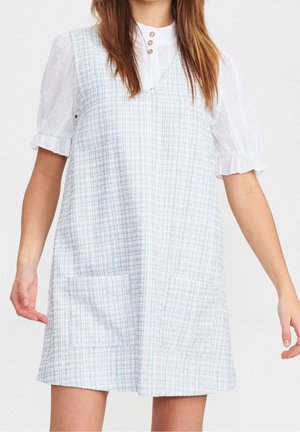 Femme portant une robe sans manches à carreaux bleu clair et blanc par-dessus un chemisier blanc à manches bouffantes avec des boutons près du col.
