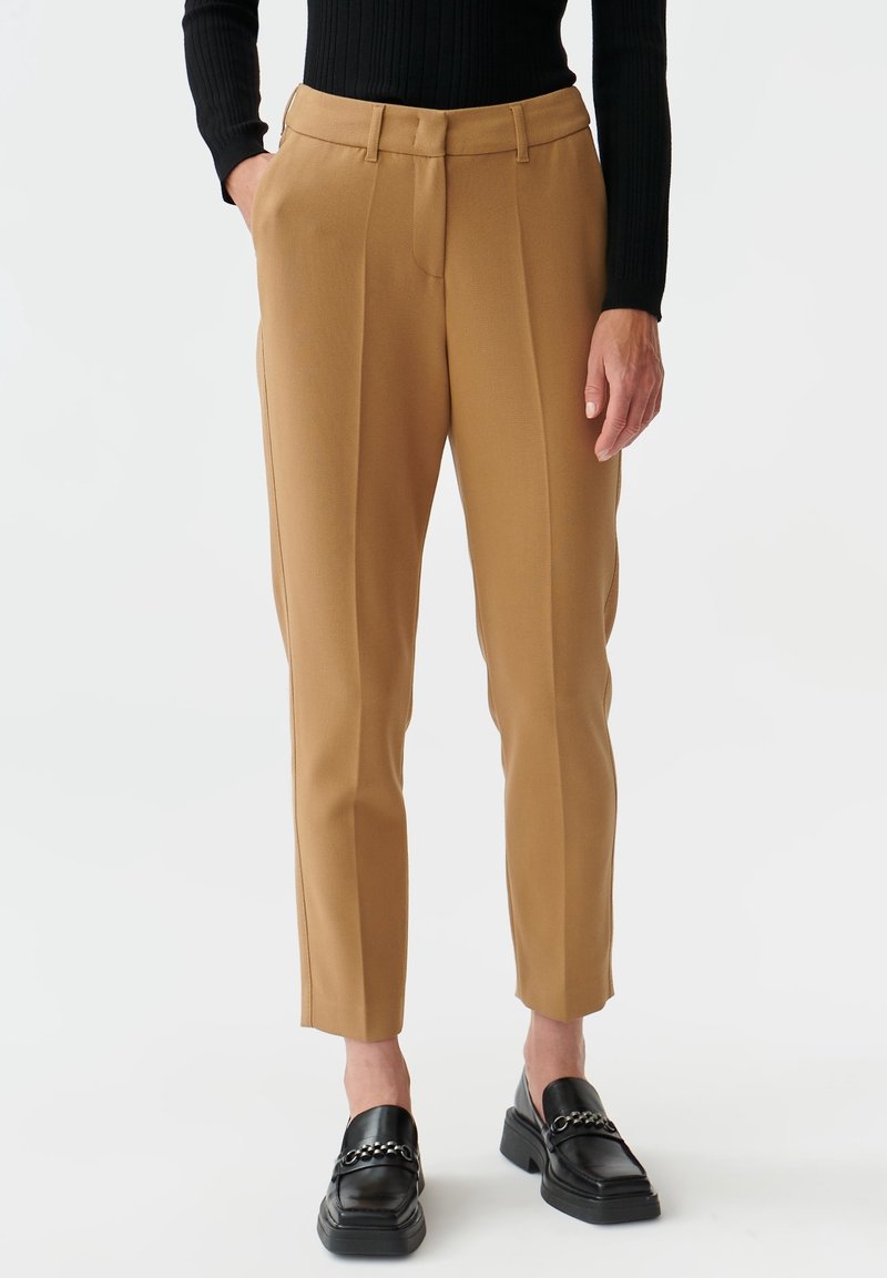 Pantalon sur mesure couleur beige en tissu lisse, doté d'une taille haute, de jambes droites et de plis repassés, assorti à des mocassins noirs.
