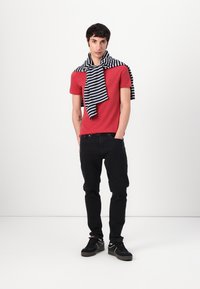 Rotes Baumwoll-T-Shirt mit kurzen Ärmeln, kombiniert mit schwarzen Hosen. Ein gestreifter Pullover in Marineblau und Weiß liegt lässig über den Schultern. Schwarze Sneaker runden das Outfit ab.