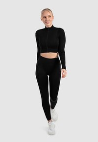 Top corto nero a coste con collo alto e maniche lunghe, abbinato a leggings neri a vita alta. Accessori includono sneaker bianche.
