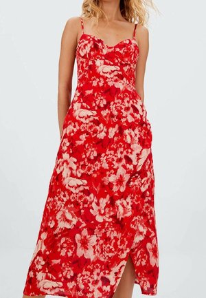 Kvinde iført en rød midi-kjole med blomsterprint, tynde stropper og en frontslids, stående foran en ensfarvet lys baggrund.