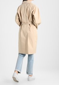 Donna con un trench coat beige con cintura, jeans blu chiaro con risvolto e sneakers bianche con plateau che si allontana su uno sfondo bianco uniforme.