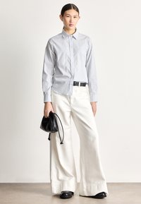 Cămașă cu dungi, cu nasturi, în albastru deschis și alb, asortată cu pantaloni largi crem. Ține o geantă mică din piele neagră.