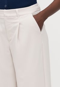 Beige wijde broek met plooien, zijzakken en een gladde textuur. Gecombineerd met een blauwe geribbelde trui, die een deel van de tailleband onthult.