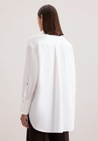 Camicia bianca a maniche lunghe con vestibilità rilassata e orlo arrotondato. Presenta polsini con bottone e una piega centrale sul retro per una maggiore libertà di movimento.