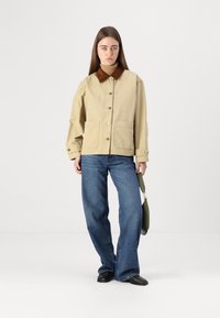 Giacca utility beige con colletto in corduroy marrone, grandi tasche frontali e bottoni dorati, abbinata a jeans a gamba larga blu e scarpe nere.