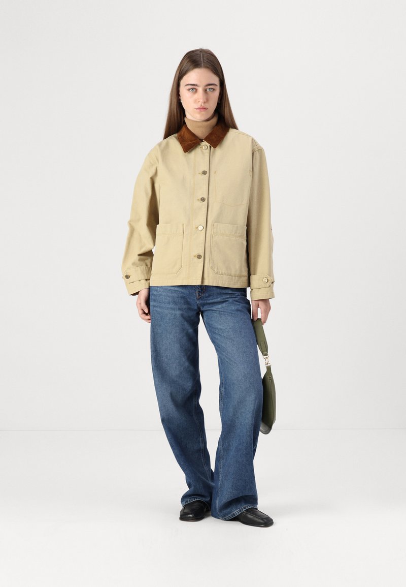 Beige Utility-Jacke mit braunem Cordkragen, großen Fronttaschen und goldenen Knopfverschlüssen, kombiniert mit blauen weit geschnittenen Jeans und schwarzen Schuhen.
