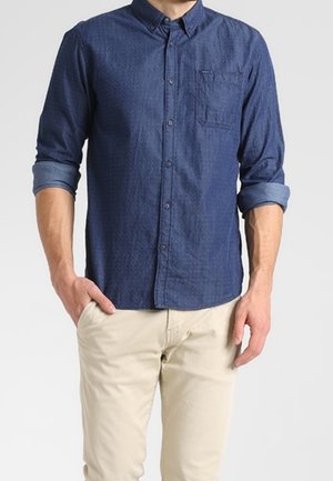 Chemise bleu marine à boutons avec col, manches retroussées et poche poitrine. Fabriquée en tissu texturé. Associée à un pantalon beige.