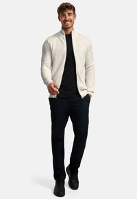 Lachende man die loopt, gekleed in een witte geribbelde trui met ritssluiting, een zwart T-shirt, zwarte broek en zwarte sneakers, tegen een effen achtergrond.