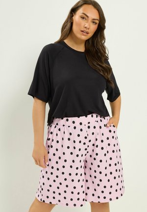Kobieta z długimi brązowymi włosami, ubrana w luźny czarny t-shirt oraz różowe spodenki do kolan w czarne groszki, ręce w kieszeniach.