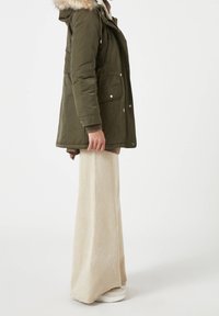 Olijfgroene parka met een nepbont capuchon, rits- en drukknoopsluiting, zijzakken, gecombineerd met beige wijde broek en witte sneakers.