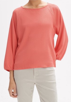 Un mannequin porte un chemisier corail texturé à encolure ronde et à manches amples trois-quarts, associé à un pantalon beige clair.