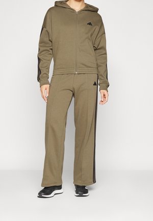 Osoba ubrana w oliwkowozieloną bluzę na zamek z kapturem oraz pasujące do niej szerokie spodnie dresowe z czarnymi paskami i czarne trampki.