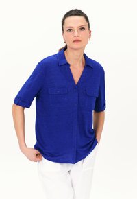 Camicetta blu a maniche corte realizzata in un tessuto texturizzato. Presenta una chiusura con bottoni frontale, due tasche sul petto e un colletto. Abbinata a pantaloni bianchi.