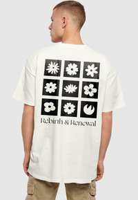 Hvid t-shirt med et sort firkantet gitter af blomsterdesigns på ryggen, med teksten "Genfødsel & Fornyelse" nederst.