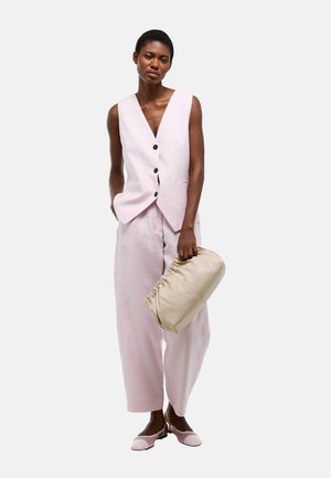 Femme portant un gilet sans manches rose clair boutonné et un pantalon large assorti, tenant une pochette beige froncée d'une main.