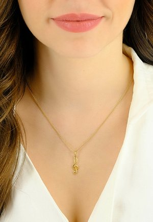 Collana tono oro con ciondolo a chiave di violino, caratterizzata da una catena sottile e un design minimalista. Indossata sopra una camicia bianca.
