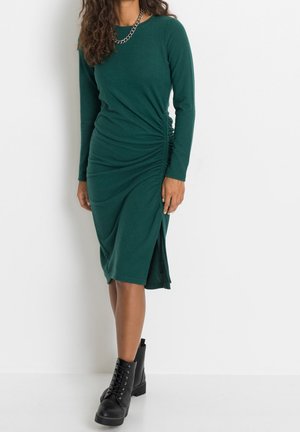 Robe longue en coton vert à manches longues, avec fronces sur les côtés et fente à l'avant, associée à des bottines noires à lacets.