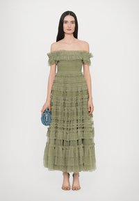 Olijfgroene off-shoulder jurk met gelaagde ruches en textuurstof, gecombineerd met een kleine blauwe clutch en nude hakken.