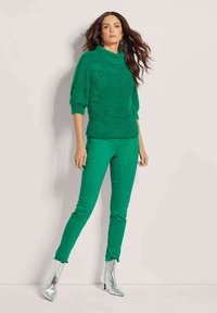 Grüner, flauschiger Pullover mit kurzen Ärmeln, kombiniert mit eng anliegenden grünen Hosen und metallisch silbernen Ankle Boots. Glatte Textur mit lockerer Passform.