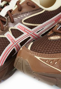 Bruna sportskor med mesh och strukturerade ytor, med rosa och vita detaljer, rundsnören och en dämpad sula.