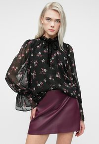 Donna bionda che indossa un blusa nera trasparente con motivo floreale e colletto arricciato, abbinata a una minigonna in pelle prugna attillata.