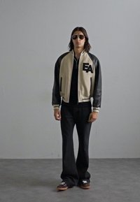 Mlad moški stoji oblečen v sončna očala, bež in črno jakno z oznako "EA" na prsih, črno majico, razširjene črne hlače in rjave čevlje.