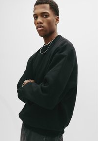 Sort sweatshirt med rund halsudskæring og lange ærmer. Blødt stof, afslappet pasform og ribbede manchetter og kant. Model iført den mod en hvid baggrund.